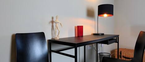 A small, modern room with a desk, two black chairs, a red vase, a wooden mannequin, and a floor lamp.