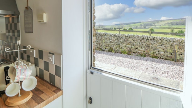 A kitchen with a stone wall view, a hanging rack with cups, and a window with a countryside view. - School House Cottage (Buxton)