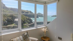 A room with a large window overlooking a coastal landscape, a chair, a small table with a vase, and a wall switch. - Penelope (Helston)