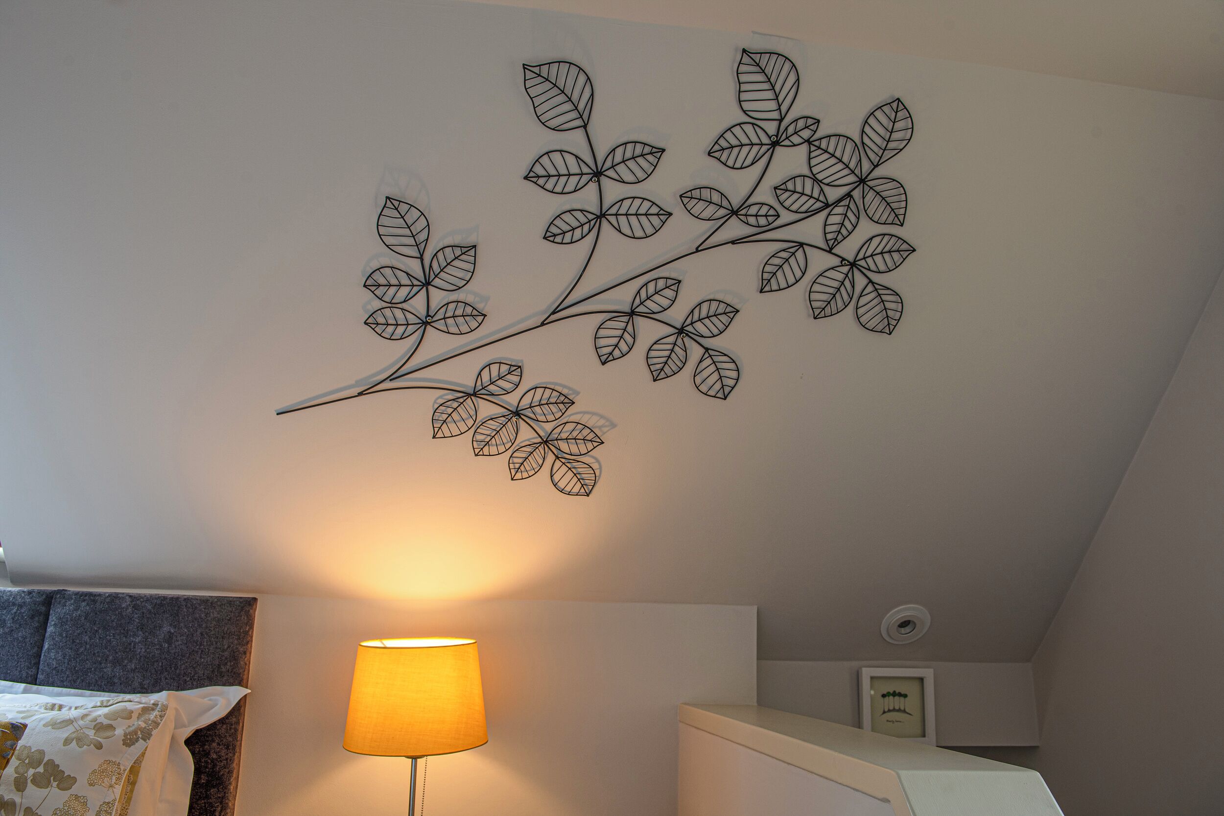 Een slaapkamer met een bed, een bedlampje en een wanddecoratie van bladeren.