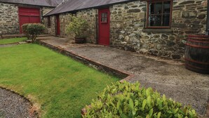 A stone building with a red door and window, a grassy yard, and a large barrel. - Preseli Hills Cottage (Fishguard)