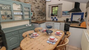 A cozy kitchen with a wooden dining table set for four, surrounded by stone walls and a blue cabinet with glass doors. - Preseli Hills Cottage (Fishguard)