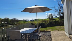 An outdoor patio with a table and chairs, a pergola with a canopy, and a view of rolling hills and a body of water in the background. - Calvey's Cottage (Newport)