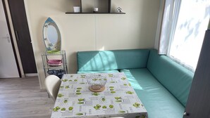 A small, cozy room with a teal sofa, a table with a cactus-patterned tablecloth, a chair, a mirror, and a shelf with decorative items. - Welcome to Lily's Mobile Home (Bernières-sur-Mer)