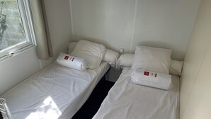Two single beds with white bedding and pillows, a window with curtains, and a metal rack. - Welcome to Lily's Mobile Home (Bernières-sur-Mer)