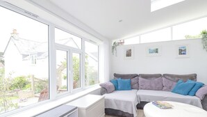 Una habitación luminosa con un ventanal grande, un sofá, un reposapiés blanco y una mesita con una revista.