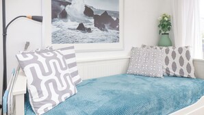 Un dormitorio con cama, almohadas, un cuadro enmarco de una costa rocosa, una lámpara y una ventana con cortinas.