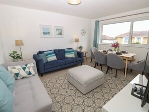 A modern living room with a sofa, ottoman, dining table, and chairs. There are two wall-mounted lamps, a clock, and framed pictures. - Sundowners (Ryde)
