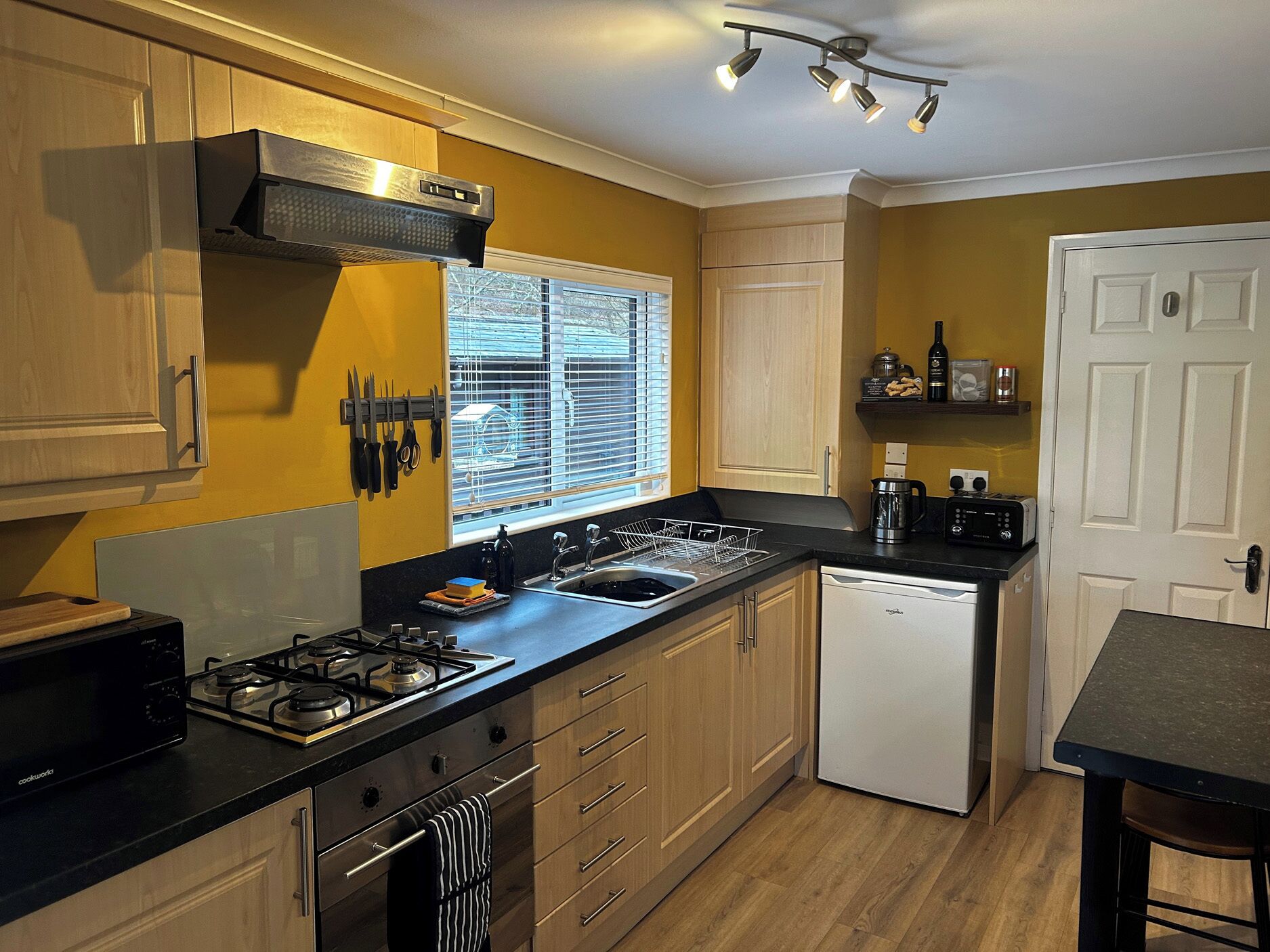 A compact kitchen with a gas stove, a dishwasher, wooden cabinets, and a window with blinds.