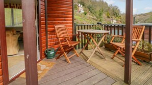 A wooden deck with two wooden chairs and a table, a green watering can, and a wooden door. - 22 Two Little Ducks (Lynton)