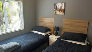 A twin room with two beds, a nightstand with a lamp, a painting on the wall, and a window with blinds. - 22 Two Little Ducks (Lynton)