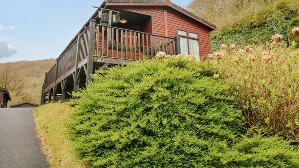 A red wooden cabin with a balcony, surrounded by lush greenery and hills. - 22 Two Little Ducks (Lynton)
