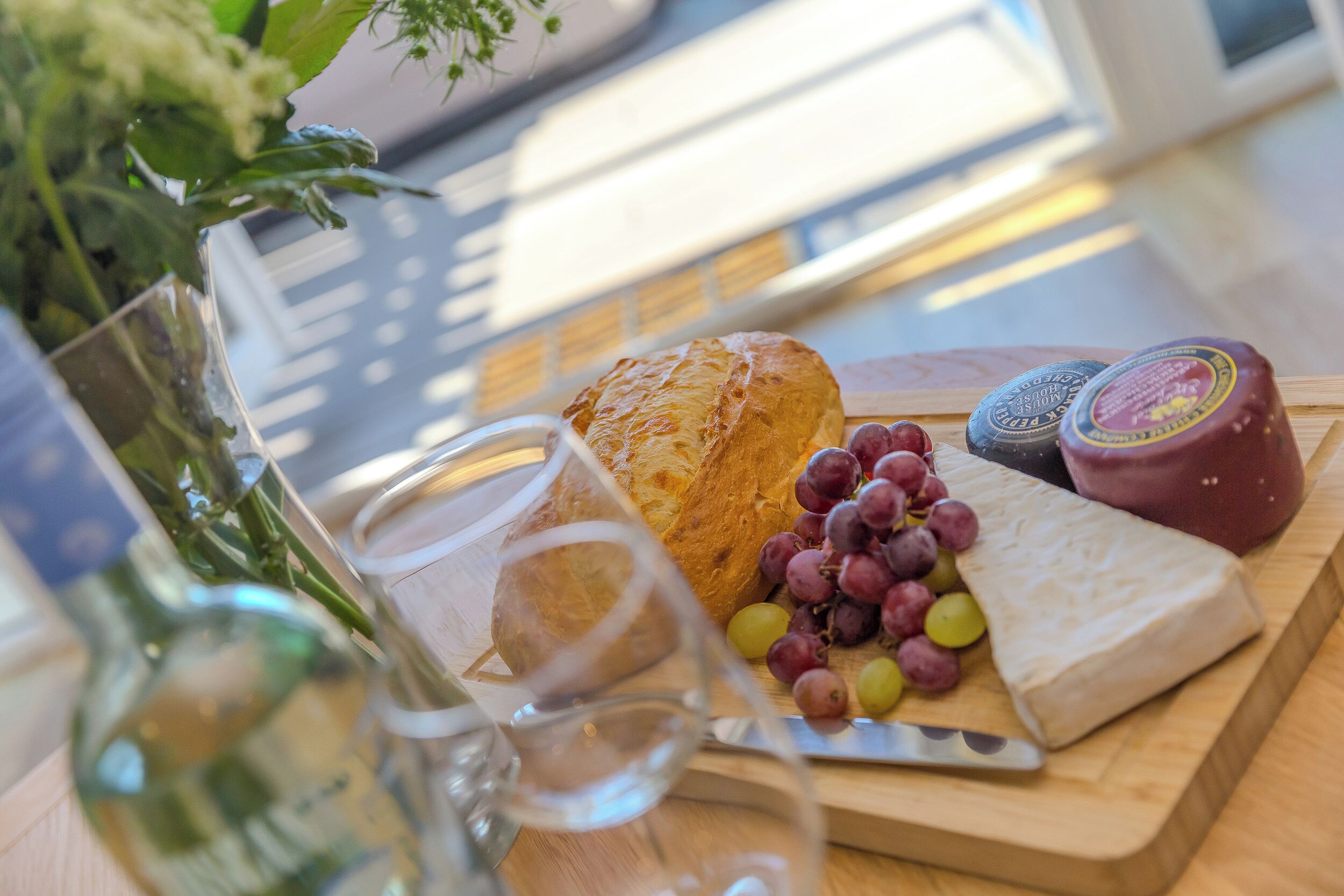 A wooden table with a cheese board, grapes, a baguette, and a bottle of wine.