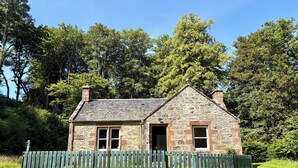 A stone cottage with a wooden fence, a grassy yard, and trees in the background.