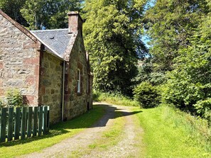A stone building with a sloped roof and a chimney, surrounded by lush greenery and a gravel path. - Gunroom Cottage (Strathpeffer)