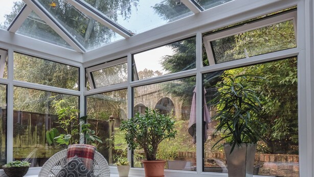 A conservatory with a glass roof, wicker furniture, potted plants, and a view of a stone bridge. - New Tower House- 10 Pear Tree Court (York)