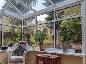 A conservatory with a glass roof, wicker furniture, potted plants, and a view of a stone bridge. - New Tower House- 10 Pear Tree Court (York)