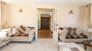 A cozy living room with two sofas, a coffee table, and a patterned ottoman. A door leads to a hallway with a wooden floor. - The Old Chapel (Dereham)