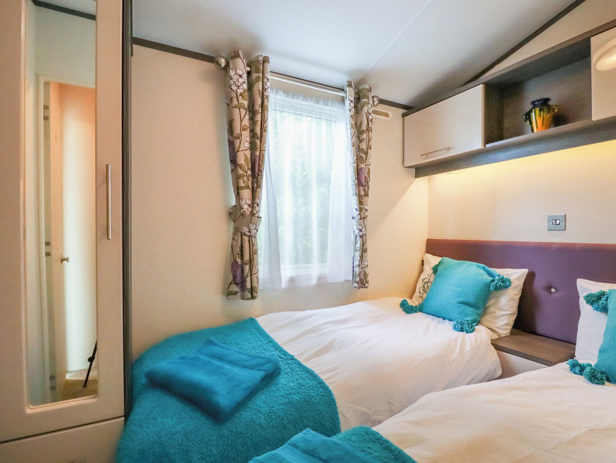 A bedroom with two beds, floral curtains, a window with sheer white curtains, and a shelf with a vase.