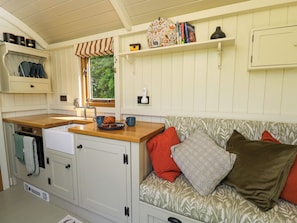 Une cuisine compacte dotée d’un plan de travail en bois, d’un évier et d’une fenêtre donnant sur un espace verdoyant. On y trouve un meuble blanc sur lequel est accrochée une serviette, ainsi qu’une petite étagère avec des livres et un objet décoratif.