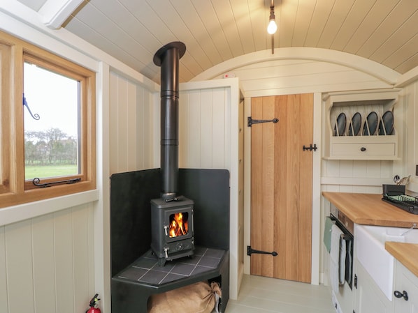 A cozy kitchen with a wood-burning stove, a window with a view, and a door leading to another room. - Spindle (Witney)