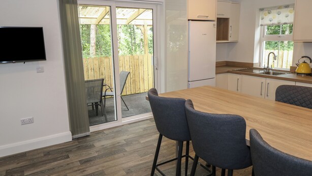 A modern kitchen with a wooden table, bar stools, a refrigerator, and a TV mounted on the wall. - Ciara's Place (Riverstown)