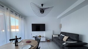 Ein modernes Wohnzimmer mit Essbereich, einem an der Wand montierten Flachbildfernseher, einem grauen Sofa mit Kissen und einer außergewöhnlichen Deckenleuchte.