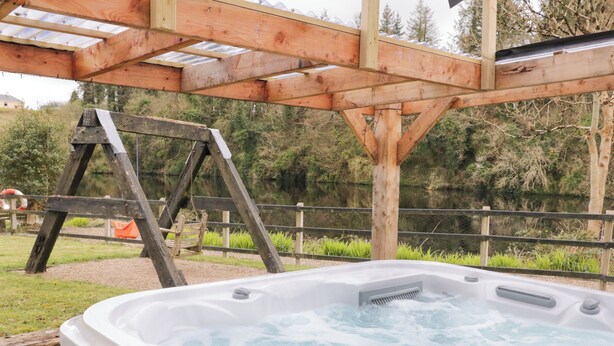 A hot tub under a wooden pergola with a wooden ladder nearby, surrounded by greenery and a body of water. - 4 Carlton Cottages (Enniskillen)