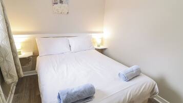 A neatly made bed with white linens, two pillows, and two rolled towels. There are bedside lamps on either side of the bed.