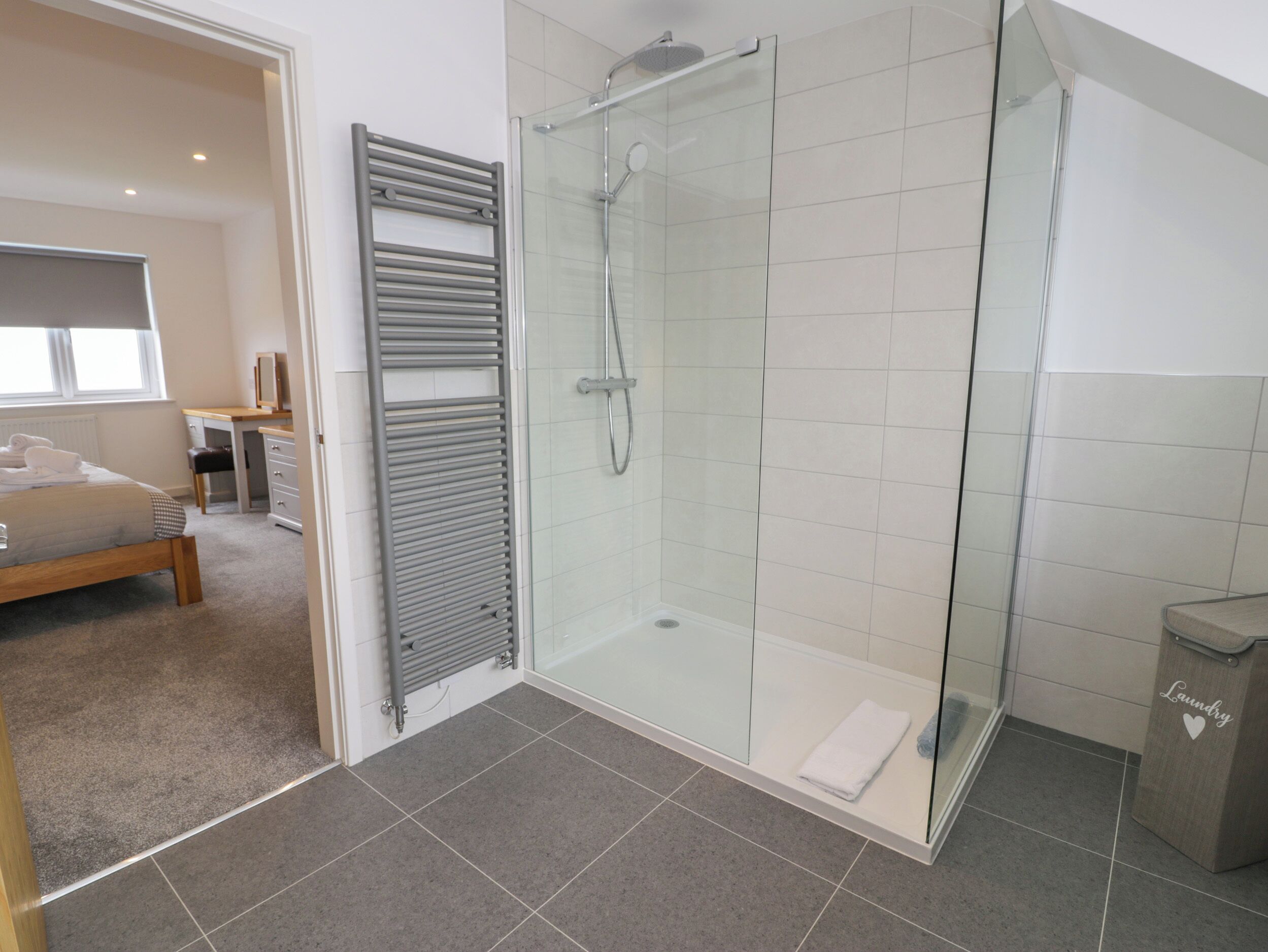 A modern bathroom with a glass-enclosed shower, a grey radiator, and a wooden bed in the adjacent room.