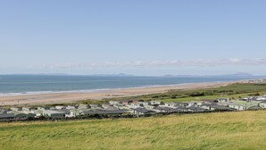 A coastal area with a row of green-roofed mobile homes along the beach, a clear sky, and distant mountains. - Melin Y Plas (Barmouth)