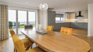 A modern dining area with a wooden table and yellow chairs, a glass vase centerpiece, and a view of the ocean through large windows. - Melin Y Plas (Barmouth)