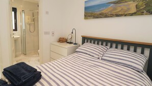 A bedroom with a bed, a nightstand, a painting of a landscape, a bathroom with a shower, and a towel on the bed. - Croyde Pebbles (Braunton)