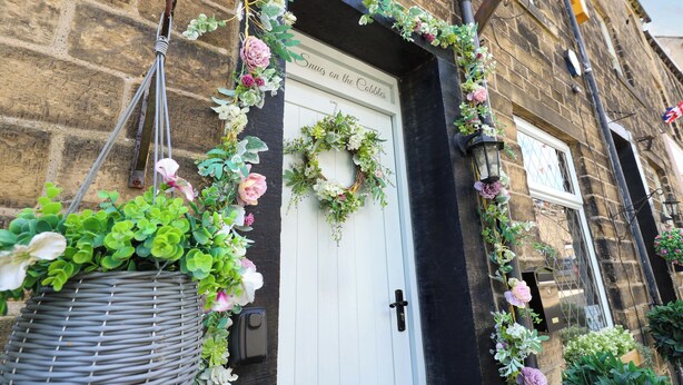 A building facade with a white door, a black awning, and a hanging basket of greenery. - The Snug on the Cobbles (Keighley)