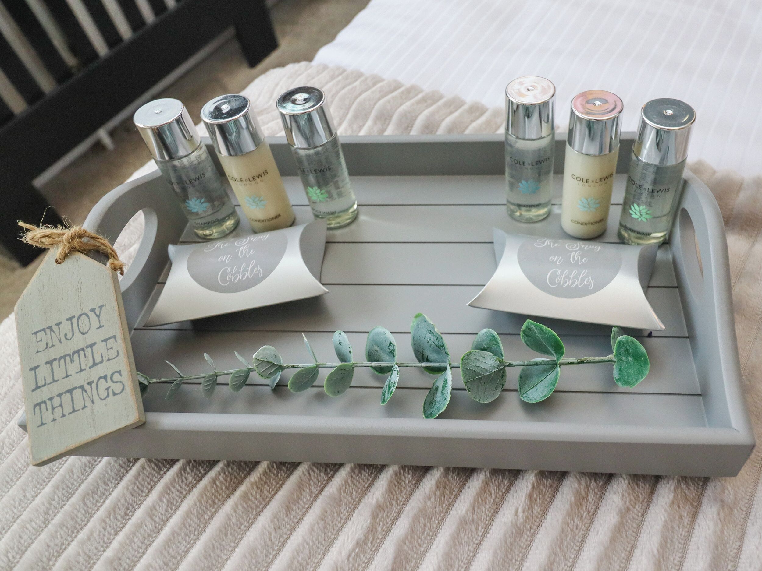Un plateau contenant six bouteilles de produits capillaires de la marque Cole + Lewin et deux étiquettes sur lesquelles est écrit « Savourez les petits plaisirs », le tout posé sur un lit recouvert d’une couverture à rayures.
