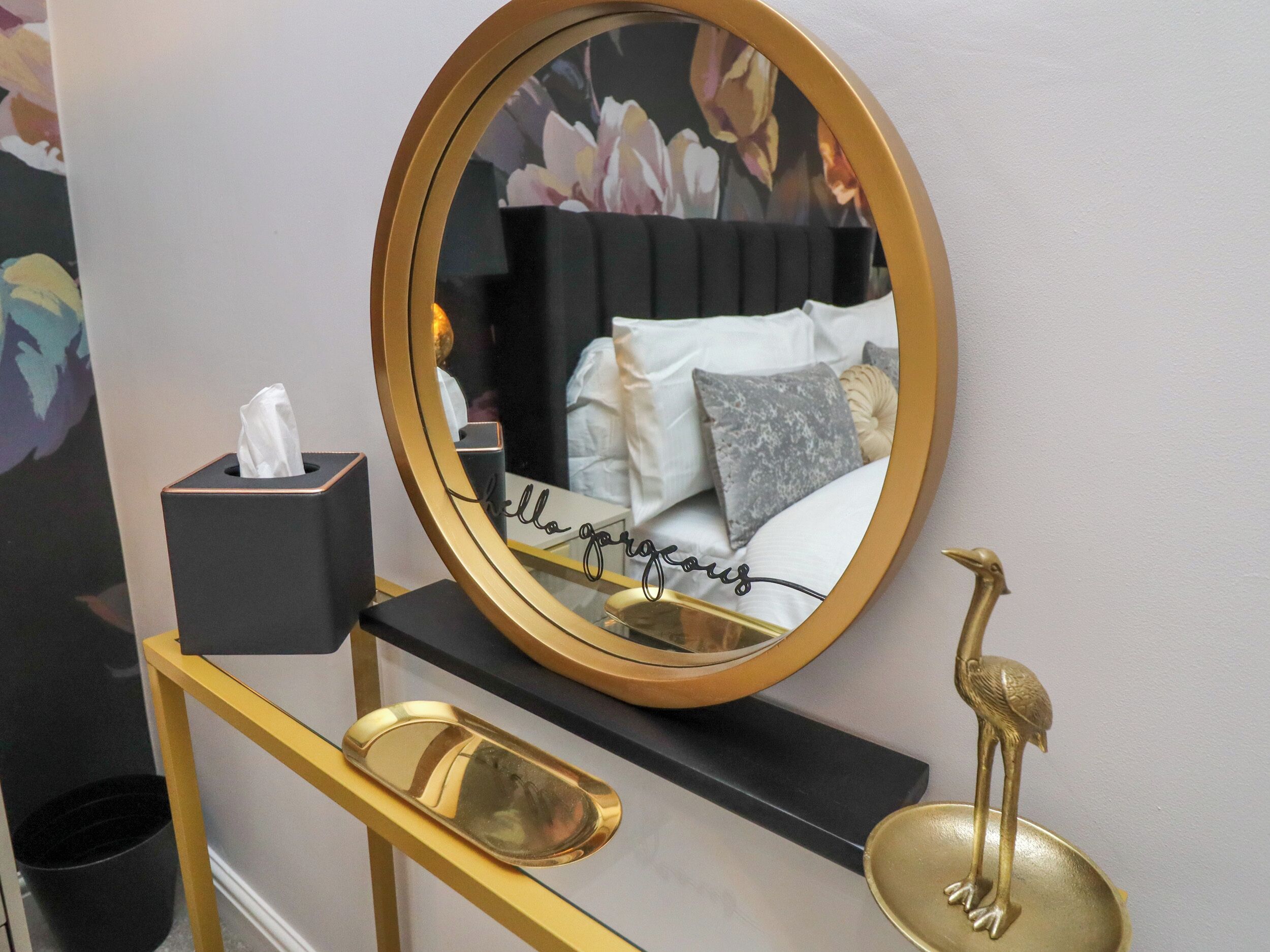 A bedroom with a round mirror, a bedside table with a tissue box, a tray, and a decorative bird.