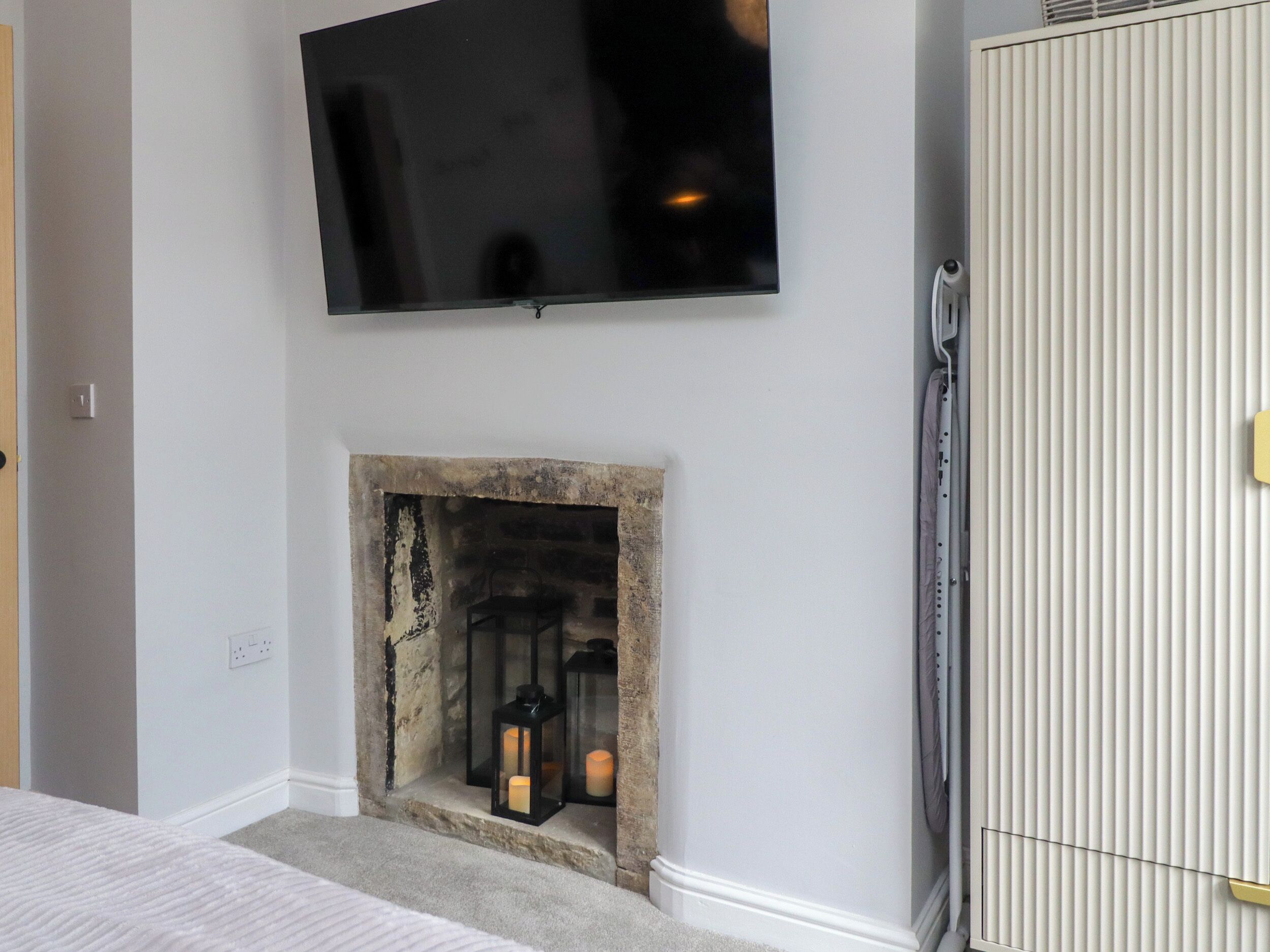 A room with a flat-screen TV mounted on the wall, a stone fireplace with candles, a radiator, and a wardrobe.