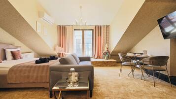 Habitación de hotel con cama, sofá, mesa de comedor con sillas, televisor montado en la pared y un amplio ventanal con cortinas.