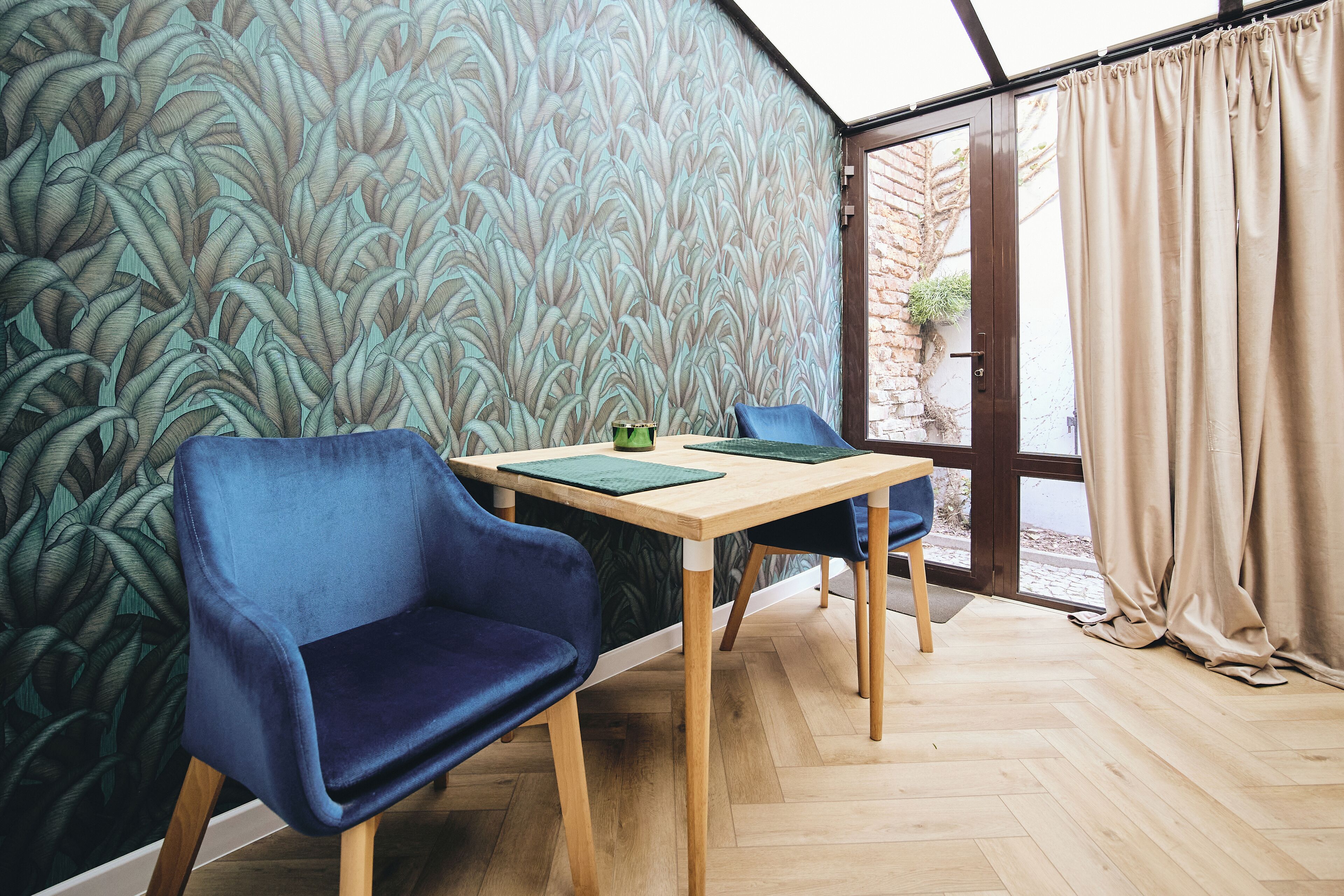 A room with a wooden herringbone floor, a table with a green placemat, two blue chairs, and a patterned wallpaper.