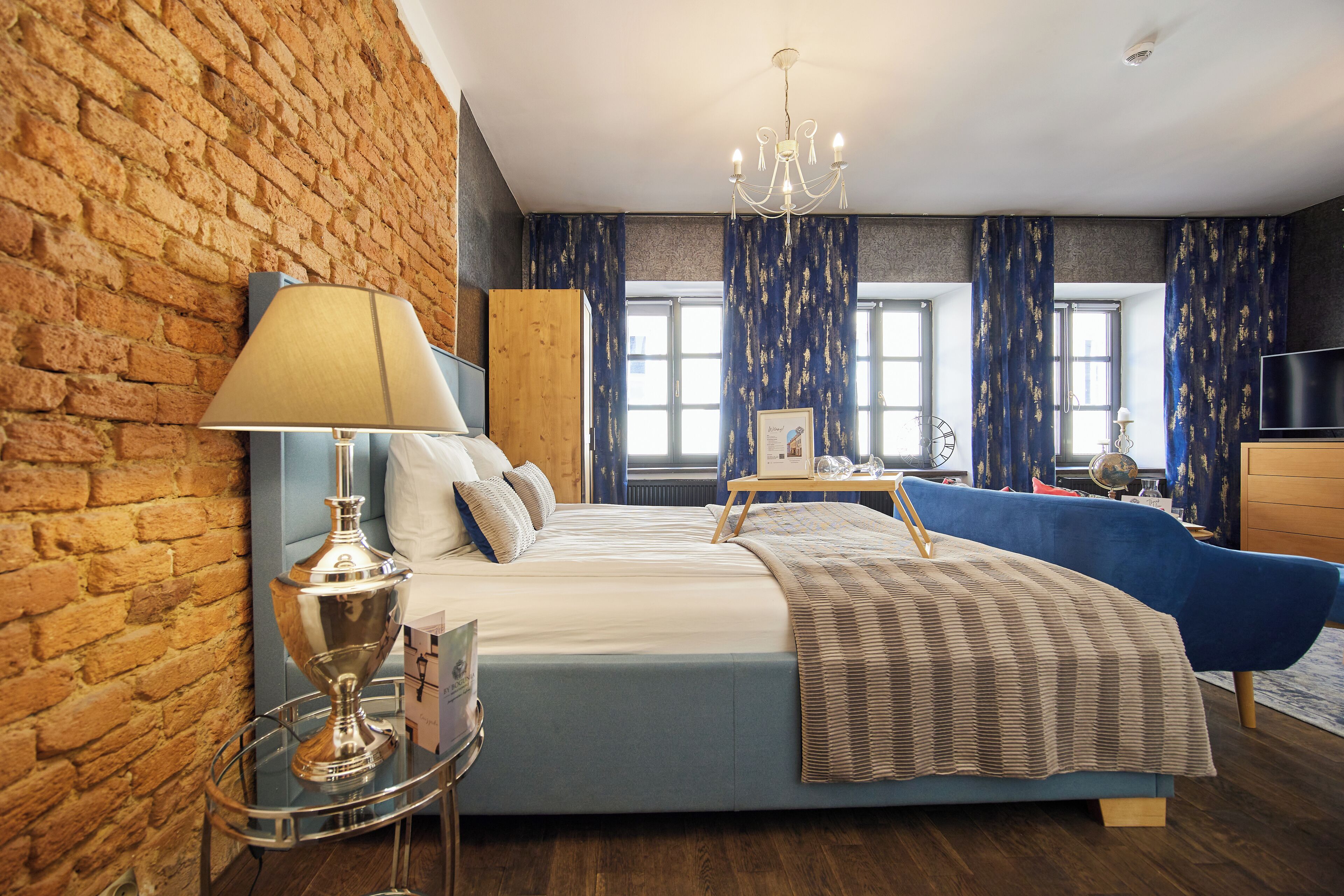Ein Hotelzimmer mit Backsteinwand, einem großen Bett, einem blauen Sessel, einem hölzernen Nachttisch und einer Chandelier.