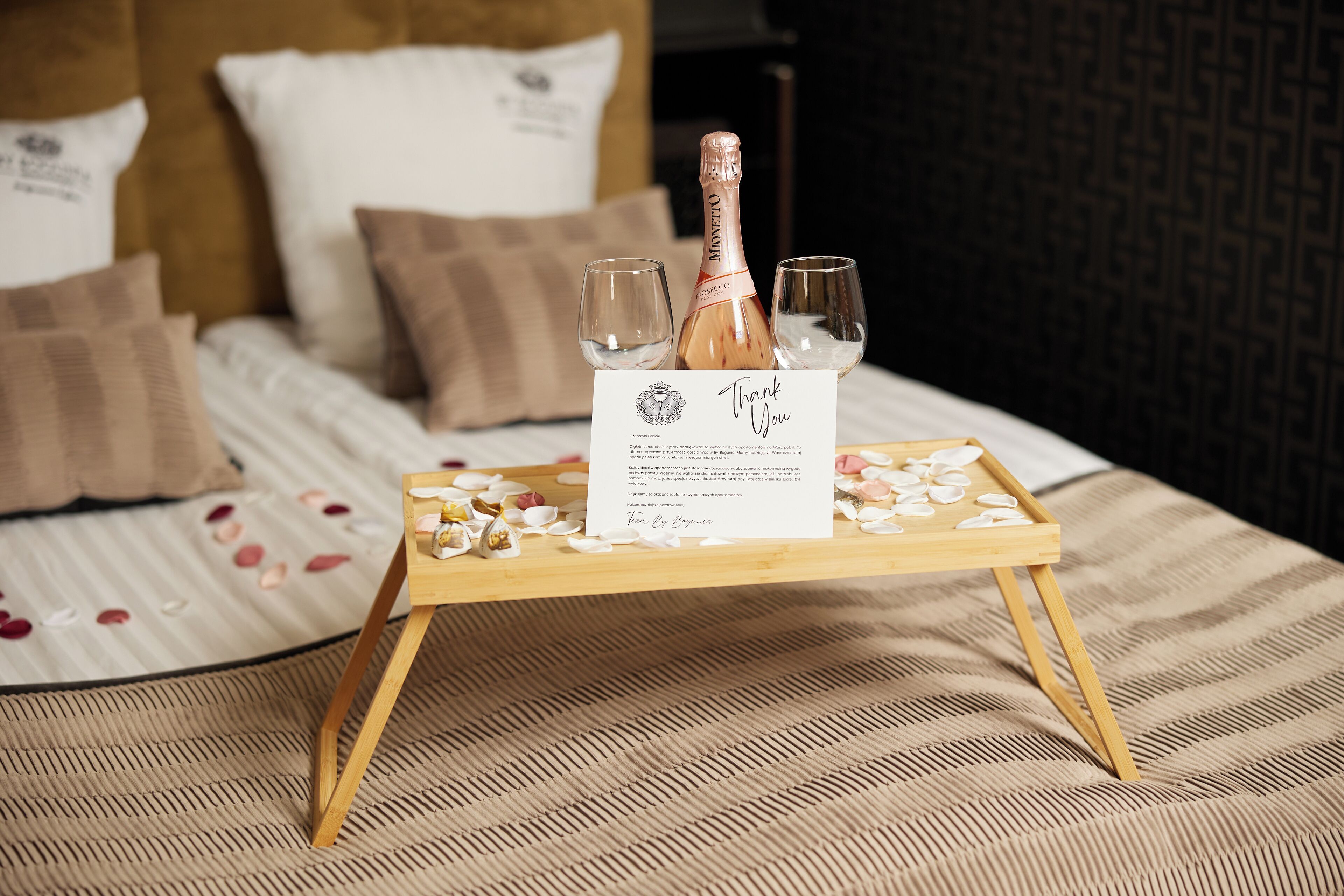 A bed with a wooden tray holding a bottle of champagne, two glasses, and a thank you card.