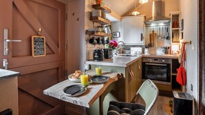 A compact kitchen with a wooden door, a chalkboard with a welcome message, a breakfast setup with croissants and juice, and a small dining area with a chair. - Penn an Vownder (The Lizard)