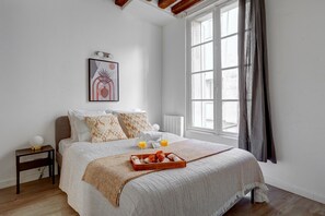 A neatly made bed with a tray of croissants and orange juice, a bedside table with a lamp, and a window with curtains. - Charming 1BR in Saint-Germain Sleeps 4 (Paris)
