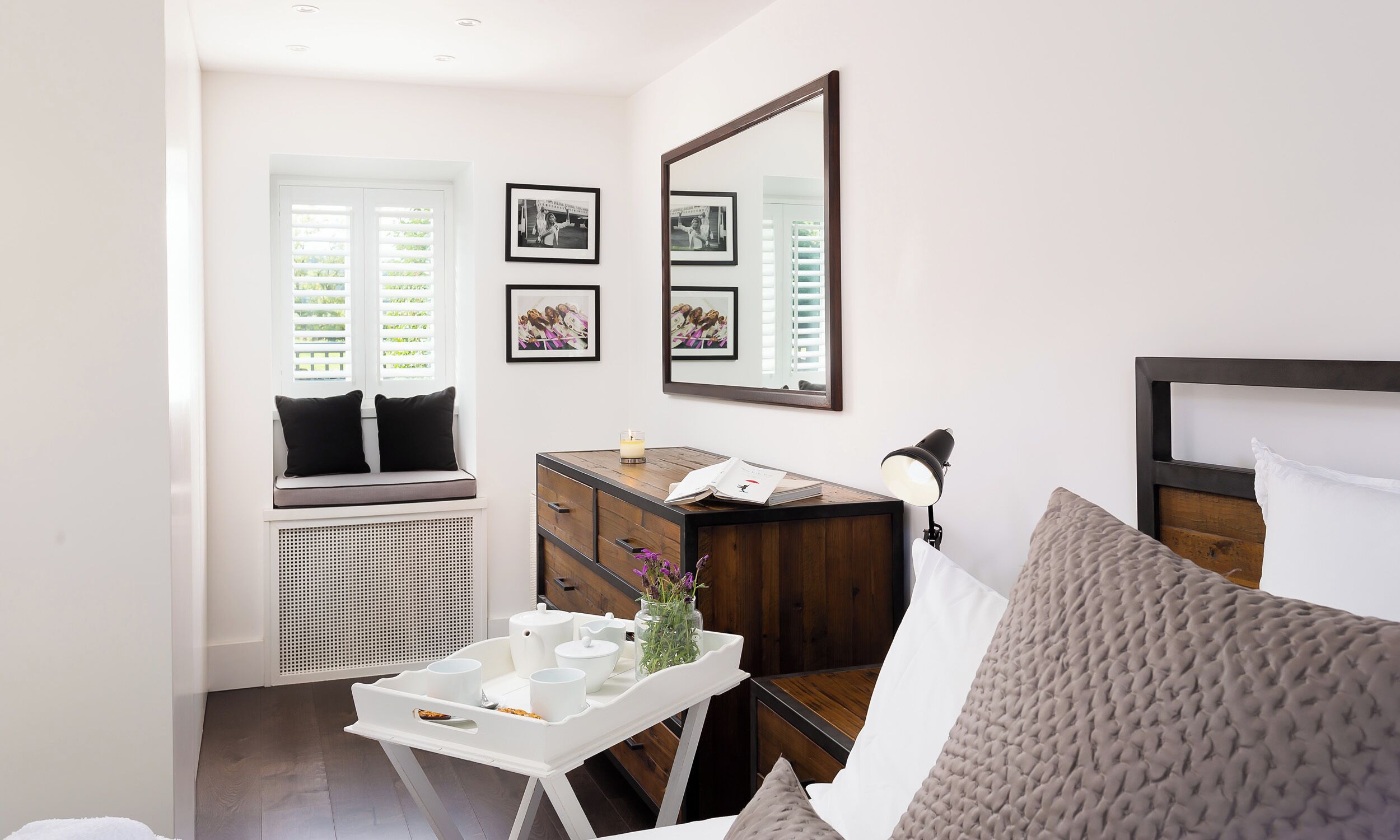 A bedroom with a bed, a bedside table, a dresser, a window with blinds, a radiator, and framed pictures on the wall.