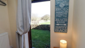 Una habitación con vista a un área verde y un muro de piedra. Un cartel en la pared dice: "Que la arena en tus zapatos siempre te recuerde que la playa es tu lugar feliz." Hay una lámpara sobre una mesa de madera junto a la pared.