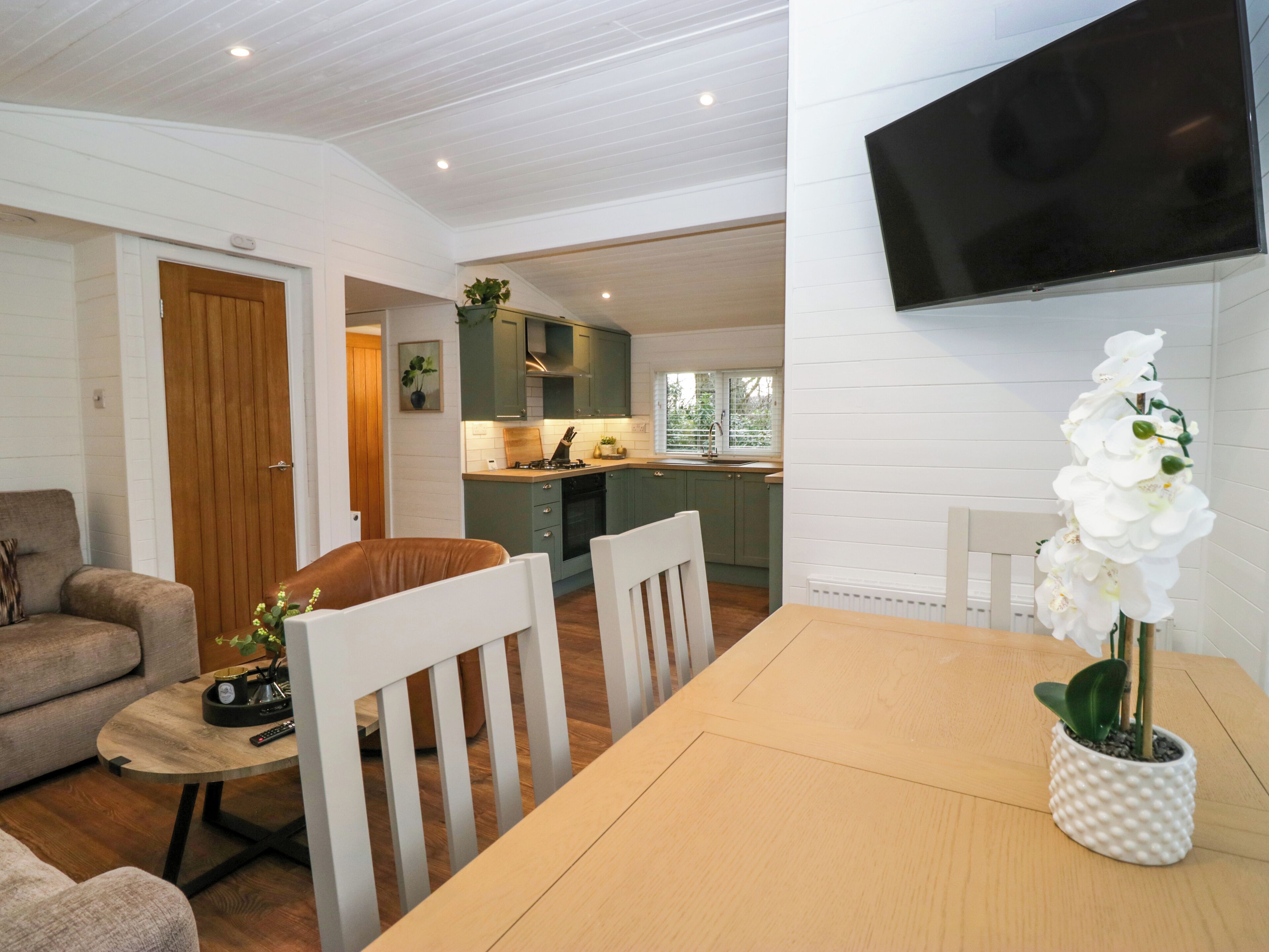 A modern living room with a sofa, a dining table with chairs, a TV mounted on the wall, and a kitchen area in the background.