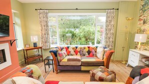A cozy living room with a colorful sofa, a small table, and a chair. There is a window with curtains, a lamp, and a bookshelf. - Esk Brae (Langholm)