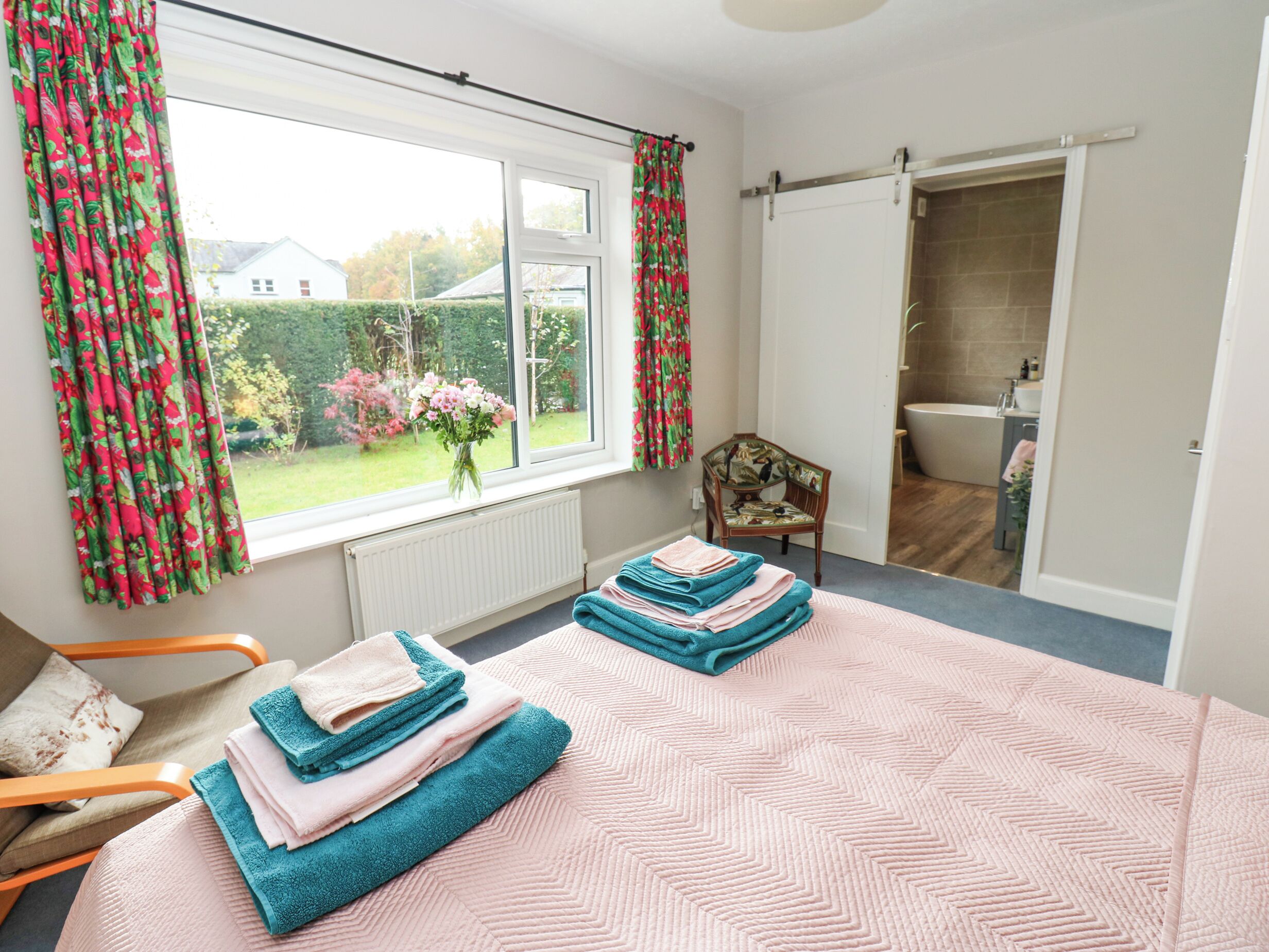 Un dormitorio con una cama grande, una silla, una ventana con cortinas y vistas a un jardín.