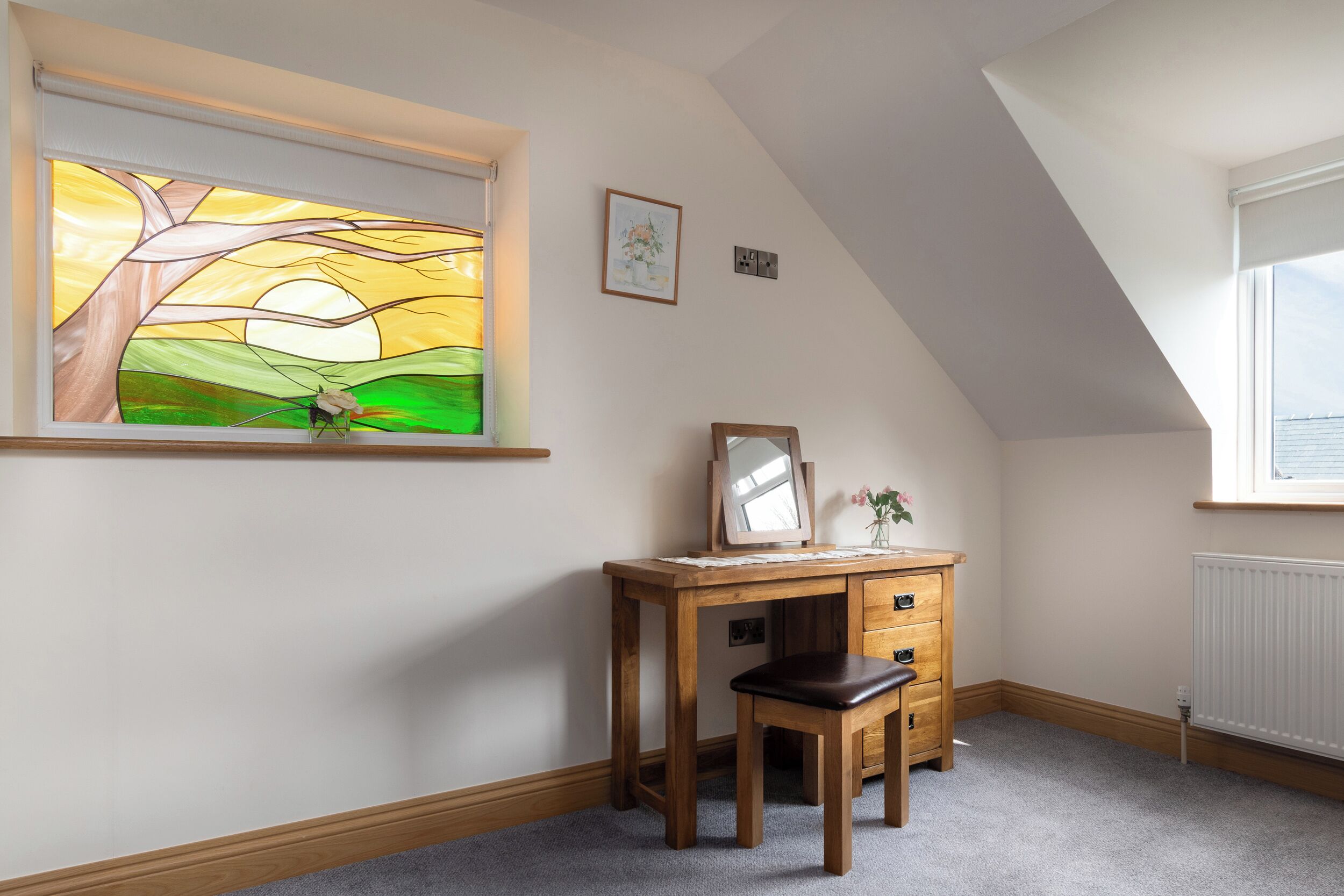 A room with a sloped ceiling, a wooden desk with drawers, a chair, a framed mirror, and a window with a stained glass view of a tree and sun.