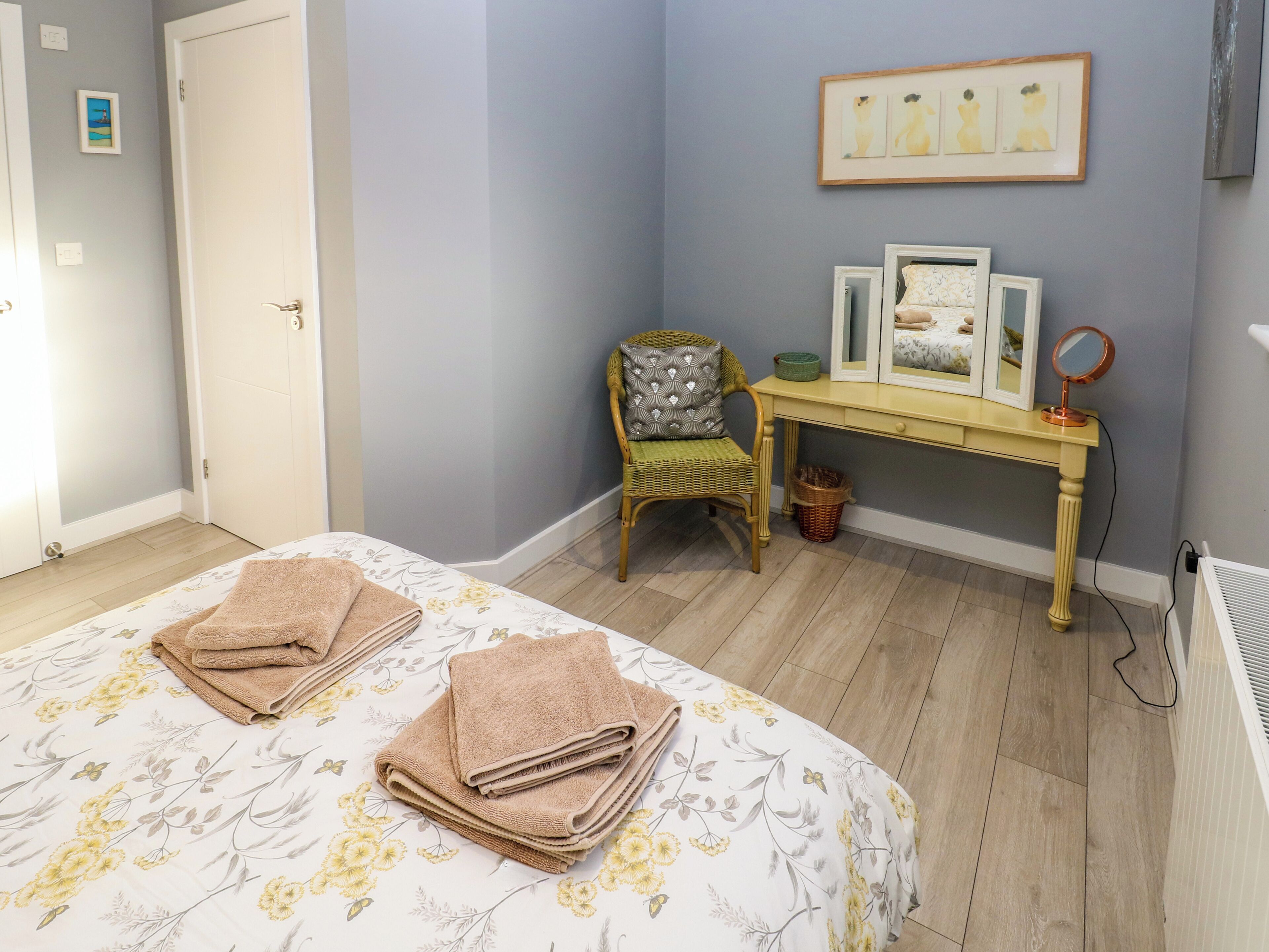 A bedroom with a bed, a chair, a vanity table with a mirror, and framed artwork on the wall.